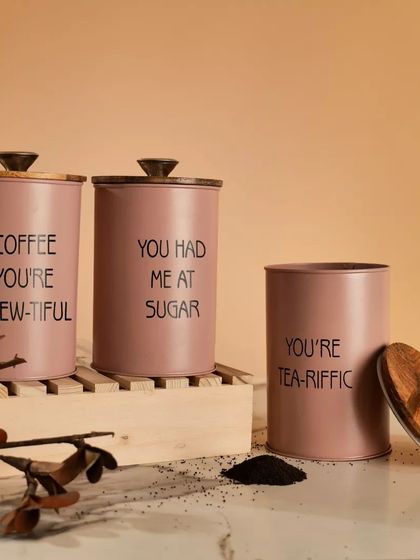 A set of pink kitchen canisters for tea, coffee, and sugar, styled with wooden elements and loose tea leaves. This shot shows the full set and suggests its use.