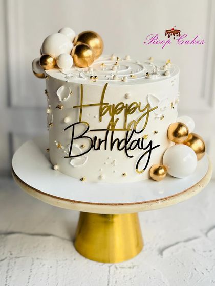 A chic and modern birthday cake in white and gold, decorated with elegant spheres and a stylish 'Happy Birthday' topper.