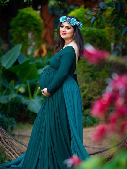 A beautiful outdoor portrait of a mom-to-be in a deep teal gown. The vibrant flowers in the foreground and the lush greenery behind her create a rich, colorful scene.