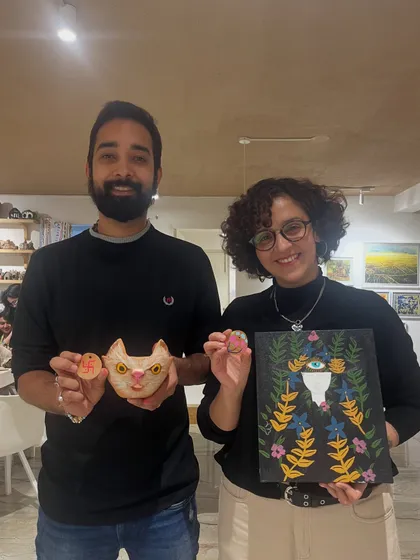 A couple poses with their unique creations, a painted cat-themed pot and a dark, floral-inspired canvas.