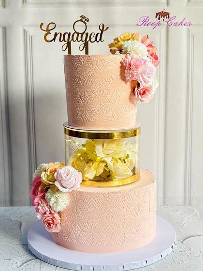 This floating tier engagement cake creates a beautiful illusion. The clear separator is filled with soft yellow flowers, connecting the two blush-pink tiers that are finished with a delicate stencil pattern and fresh floral arrangements.