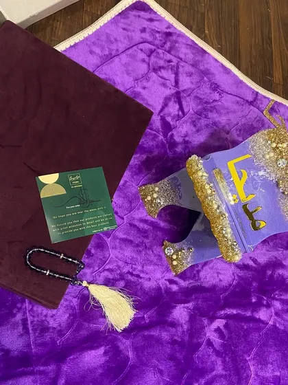 This personalized gift set features a vibrant purple prayer mat paired with a custom resin Quran holder. The holder is decorated with gold glitter and pearls, and the set includes a matching tasbeeh.