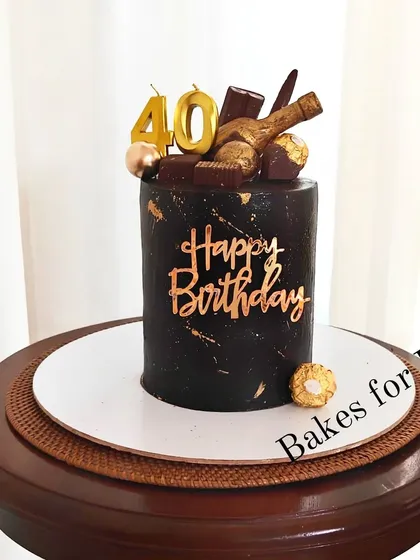 A sleek, all-black cake for a 40th birthday. This modern design is topped with a miniature chocolate bottle, Ferrero Rocher chocolates, and gold number candles.