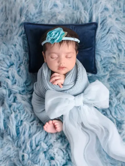 A beautifully swaddled newborn girl sleeping on a soft blue fur rug. The delicate blue floral headband and large bow add a touch of elegance to this peaceful portrait.