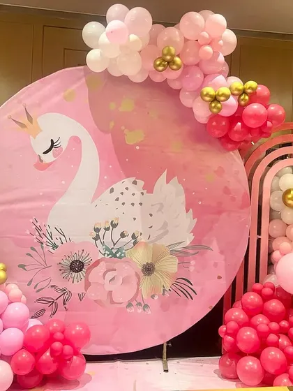 A simple and elegant swan-themed Aqeeqah ceremony. The decor features a large circular backdrop with a graceful swan graphic, surrounded by a pink, white, and gold balloon garland.