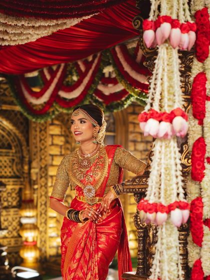 A close-up of the bride against the traditional mandap, highlighting the detailed floral work with lotus bud hangings and classic red and white garlands.