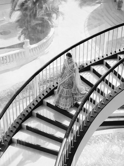 A high-angle black and white shot of the bride descending a grand, curved staircase. The graphic lines of the stairs and her flowing lehenga create a dramatic and elegant composition.