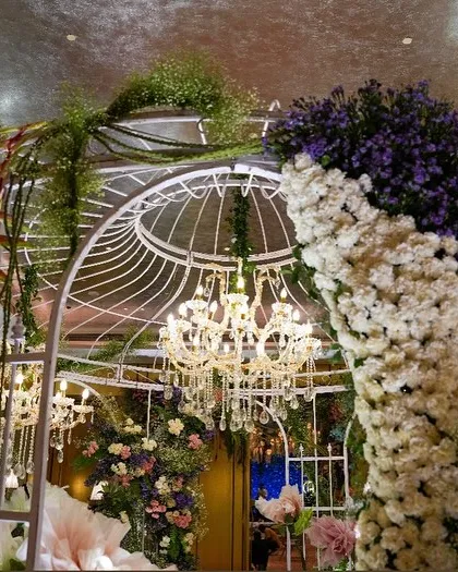 An enchanting floral birdcage structure, forming a magical entrance or photo spot. The cage is adorned with cascading white and purple flowers and houses a glittering chandelier inside.