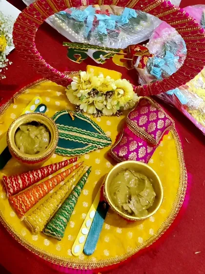 A complete Mehendi platter setup, filled with fresh henna paste in bowls, colorful decorative cones, and other traditional elements. The platter is decorated with a yellow floral gajra for the bride.