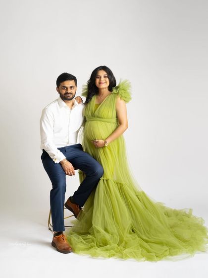 A bright and happy couple's portrait against a clean white background. The mother-to-be shines in a lime green tulle gown while her partner sits beside her, both smiling for the camera.
