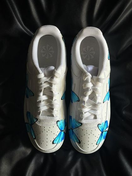 A pair of Nike Court Vision sneakers covered in delicate, hand-painted blue butterflies.
