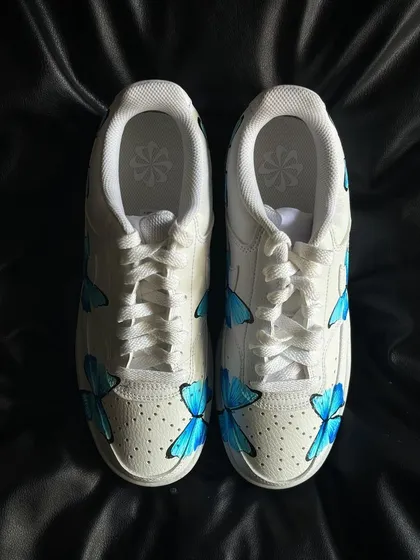 A pair of Nike Court Vision sneakers covered in delicate, hand-painted blue butterflies.