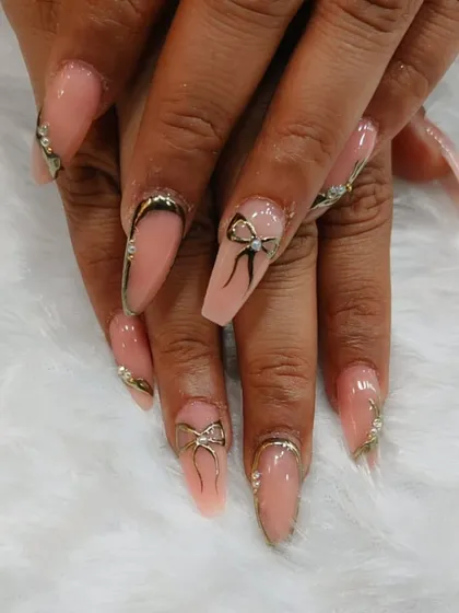 Elegant coffin-shaped nails with a nude base. The design is enhanced with delicate hand-painted black bows, gold chrome outlines, and scattered glitter for a touch of class.