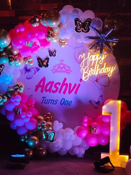 A butterfly-themed first birthday for "Aashvi," featuring a custom backdrop, a "1" marquee number, and a vibrant pink and gold balloon arch.