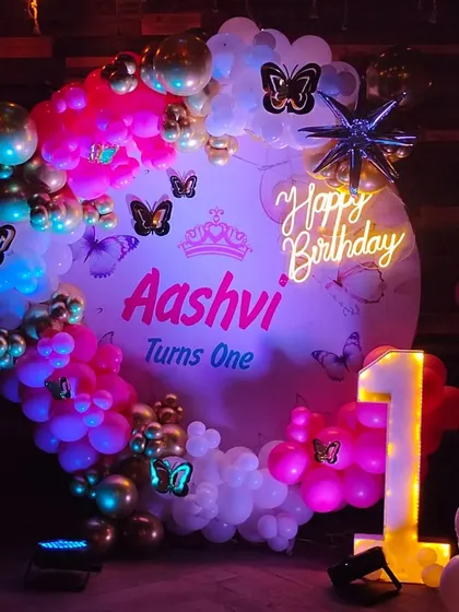 A butterfly-themed first birthday for "Aashvi," featuring a custom backdrop, a "1" marquee number, and a vibrant pink and gold balloon arch.