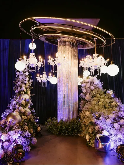 A futuristic entrance installation for a Sangeet, featuring a crystal fringe column, suspended chandeliers, and glowing orbs, all surrounded by clouds of white baby's breath.