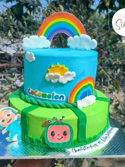 Another bright and happy Cocomelon cake. This two-tier design features a sunny sky on top and a grassy field below, with edible cutouts of the beloved characters.