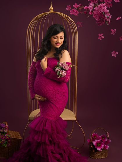 A portrait of quiet elegance. The mother-to-be, in a rich magenta gown, is seated in a golden birdcage chair, surrounded by falling petals, creating a romantic and graceful image.