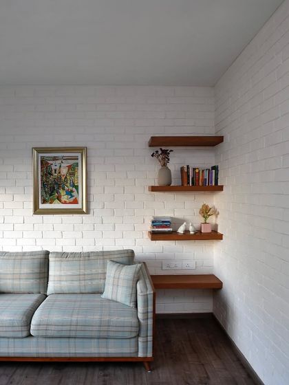A quiet corner in a home, with a comfortable plaid sofa against a white-washed brick wall. The simple floating wooden shelves provide a space for books and personal objects, creating a perfect reading nook.