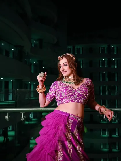 A candid shot of the anchor on a balcony at night. The ambient light from the building creates a beautiful, modern backdrop that contrasts with her traditional, vibrant lehenga.