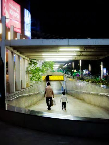 A father and child walking into a subway entrance at night. The leading lines and bright lighting create a compelling urban portrait that tells a story of guidance and togetherness.