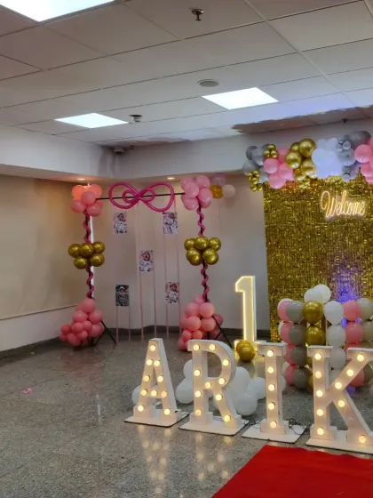 A glamorous first birthday party in a banquet hall. This decor features a gold shimmer wall, large light-up letters for the name 'ARIKA', a marquee number '1', and pink and gold balloon arrangements.