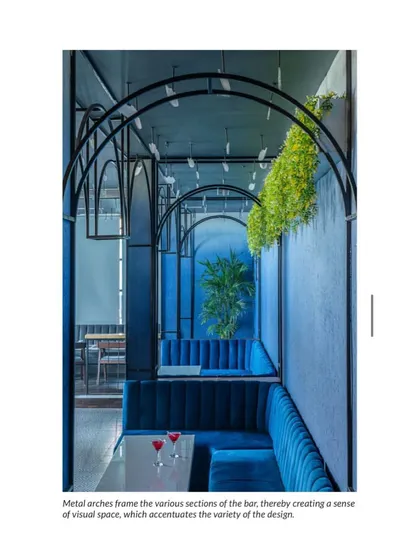 Metal arches frame the various sections of the Ombré bar, creating a sense of visual space and rhythm. The deep blue velvet booths and hanging greenery add to the moody, luxurious atmosphere.