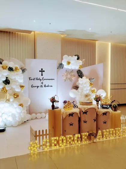 For George and Andrea's First Holy Communion, we designed a sophisticated and modern stage. The decor combines clean white panels, elegant balloon clouds, and touches of gold, with personalized marquee lights to make the day truly special and blessed.