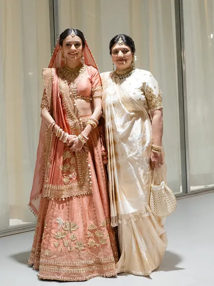 The bride with her mother, both looking radiant. The mother's classic ivory saree provides a beautiful, elegant contrast to the bride's soft pink lehenga.