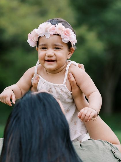 A mother lifting her smiling baby girl, crowned with flowers. A sweet and joyful moment captured in a beautiful green setting.