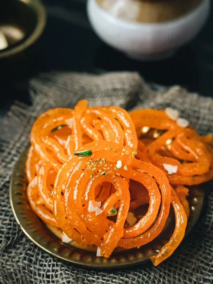 A close-up of hot, crispy jalebis on a brass plate. The dark, rustic styling makes the bright orange of the jalebis stand out, emphasizing their crunchy, syrupy goodness.