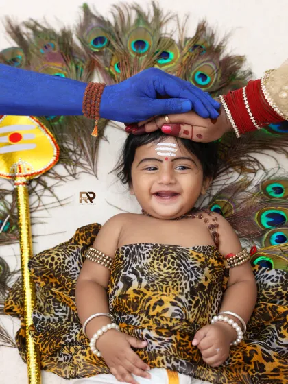 A unique and heartwarming photo celebrating Lord Shiva. A smiling baby is blessed by his parents, whose hands are painted to represent Shiva and Parvati, against a backdrop of peacock feathers.
