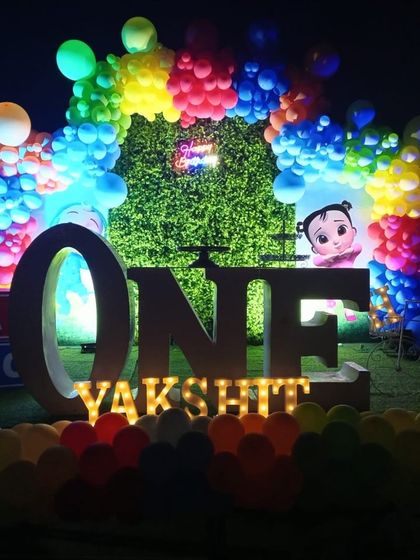 A vibrant Cocomelon first birthday setup for an outdoor night party, using a grass backdrop, rainbow balloons, and large illuminated letters.