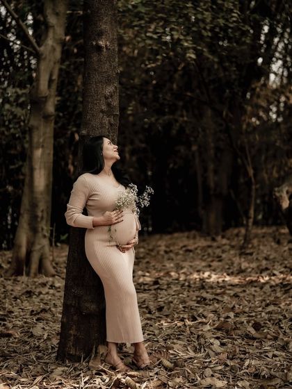 A wide shot of the mom-to-be leaning against a tree in the forest, looking up with a hopeful expression. The image captures a sense of scale and wonder.