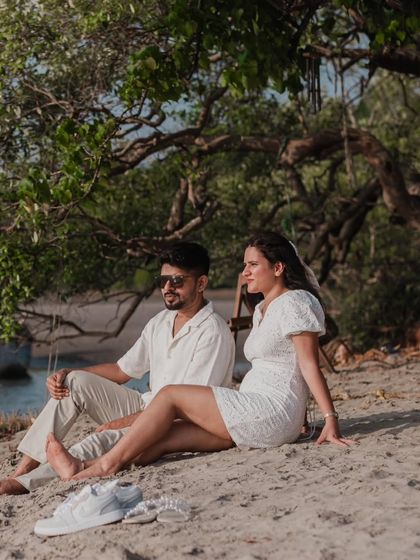 A relaxed, candid moment of the couple sitting on the sand under a tree. It feels natural and unposed, like they are just enjoying a quiet moment together.