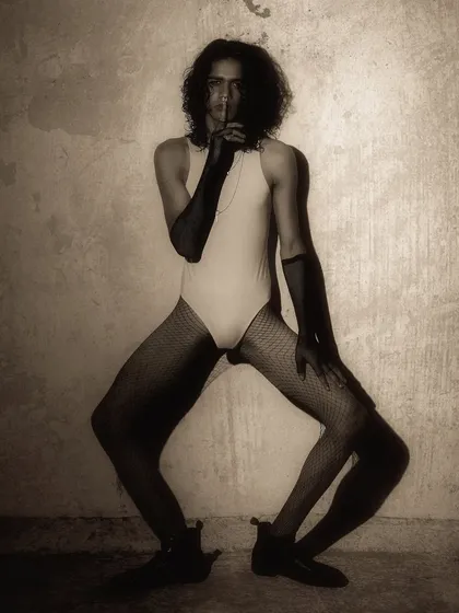 A full-body, high-fashion pose that is both angular and expressive. The lighting is dramatic, carving out the model's form against the textured wall.