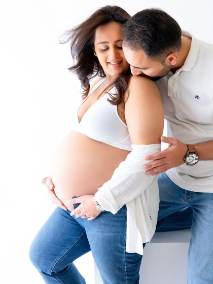 A tender moment captured between an expecting couple. The partner's gentle kiss on her shoulder adds a layer of intimacy to this casual, white-background maternity shoot.