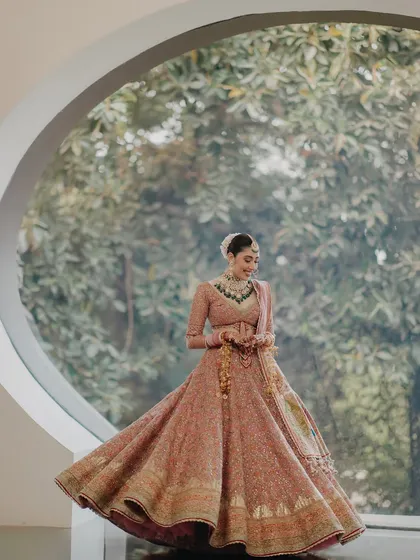 A stunning portrait of Ragini framed by a large circular window. The natural light and the lush green background create a beautiful contrast with her pink lehenga, resulting in a breathtaking shot.