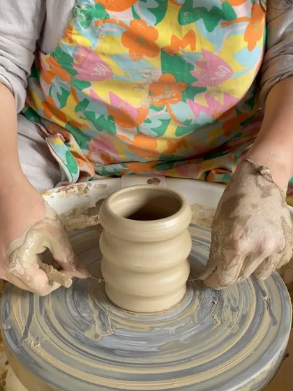 A student's hands covered in clay as she works on a piece at the wheel, capturing the tactile nature of pottery.