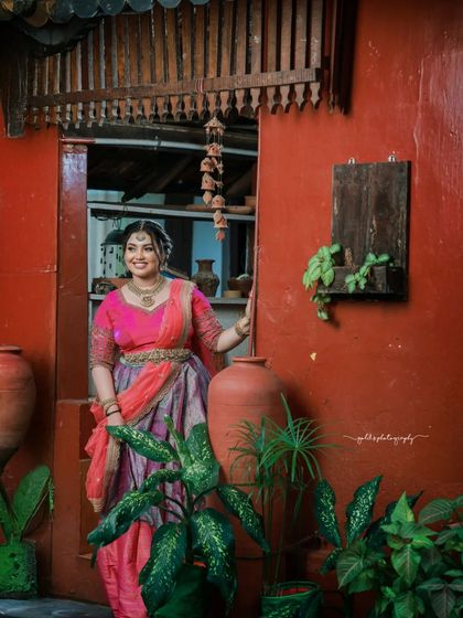 The bride stands in a traditional courtyard, with the red walls and green plants providing a colorful and rustic backdrop.
