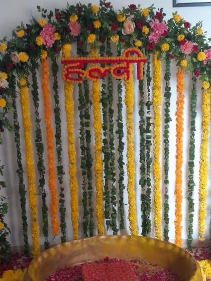 A classic Haldi ceremony arch with a mix of colorful flowers and hanging garlands. The large brass urli in the center is filled with rose petals, ready for the rituals.