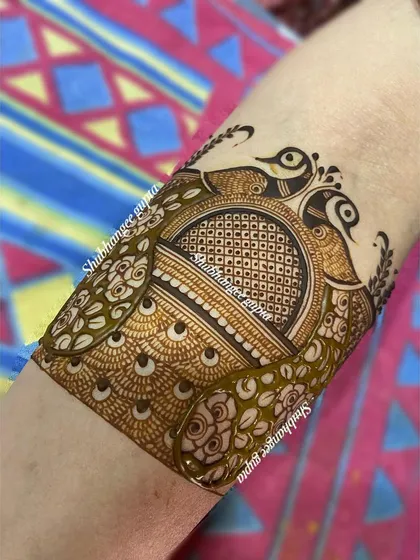 A beautiful arm band design featuring two peacocks facing each other. The body of the peacocks is filled with a fine checkerboard pattern, adding texture and detail.