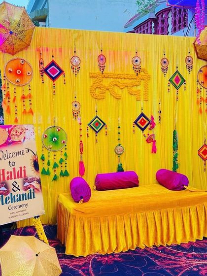 An outdoor Haldi and Mehendi setup with a bright yellow backdrop and traditional hangings. I included a custom welcome sign on an easel to greet guests, and the seating area is accented with Rajasthani umbrellas for a festive feel.