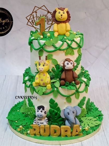 A classic two-tier jungle-themed cake for a first birthday. The cake is covered in fondant vines and leaves and features cute fondant animals like a lion, giraffe, monkey, and zebra.