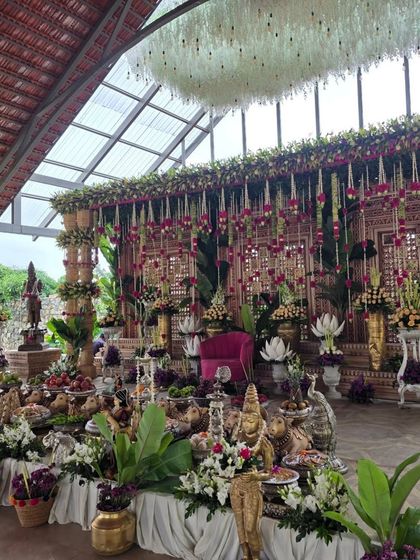 A grand ceremony setup inside a venue with a glass ceiling, giving it an outdoor feel. The decor is rich with purple and white flowers, banana leaf pillars, and a large offering table.