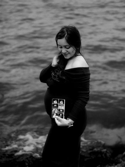 A black and white portrait of the mom-to-be by the water, looking down at her sonogram pictures. The waves and her quiet expression create a moody and emotional scene.