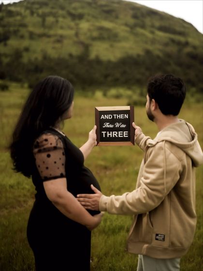 A beautiful pose from behind, with the couple holding the "And Then There Were Three" sign towards the scenic hills. His hand on her bump adds a layer of connection and tenderness.