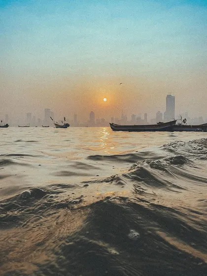 A low-angle shot from the water level at sunset, with fishing boats and the city skyline in the distance. This captures the life of the Koli fishermen.