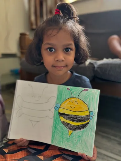 A junior KG student holds up her cheerful bee drawing. This exercise helps little ones practice drawing basic shapes and patterns, like the stripes on the bee, while creating a cute character.
