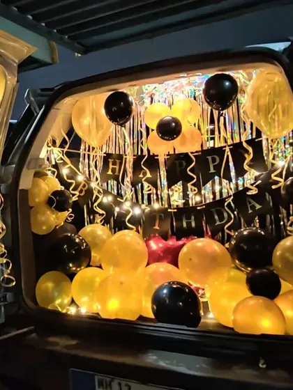 A birthday car trunk surprise illuminated for a nighttime reveal. This setup uses yellow and black balloons, a "Happy Birthday" banner, and string lights to create a magical effect in the dark.